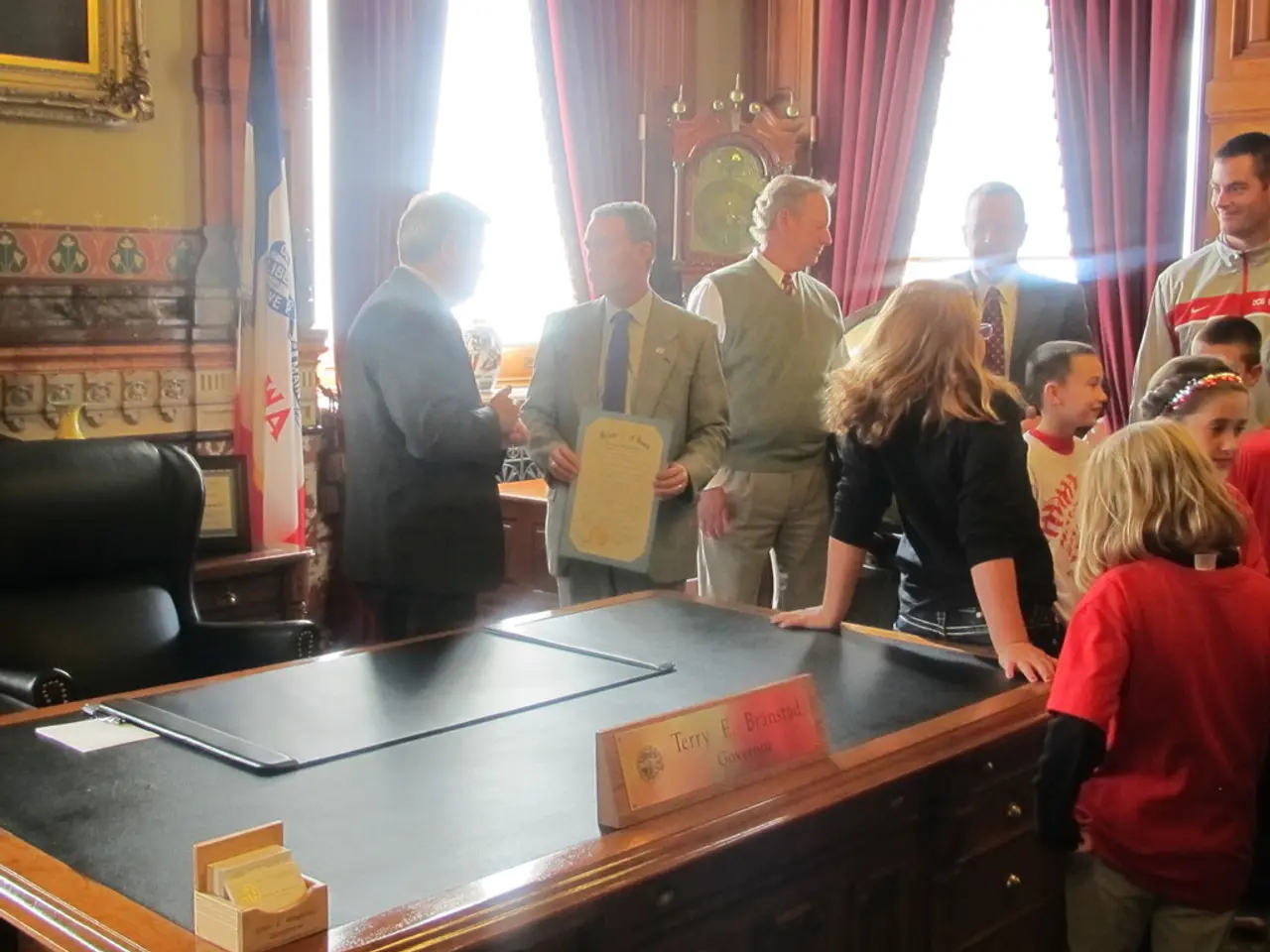 In the image there are few people stood talking and smiling, in the back of them there is a table....