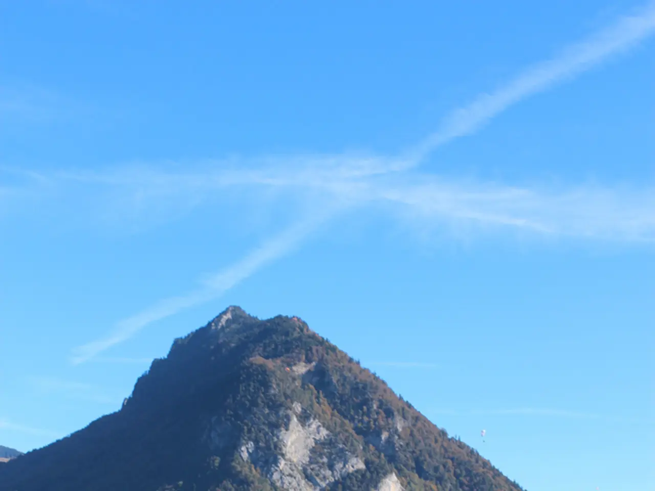 In this image we can see sky with clouds, hills, trees, flags, flag posts, buildings and water.