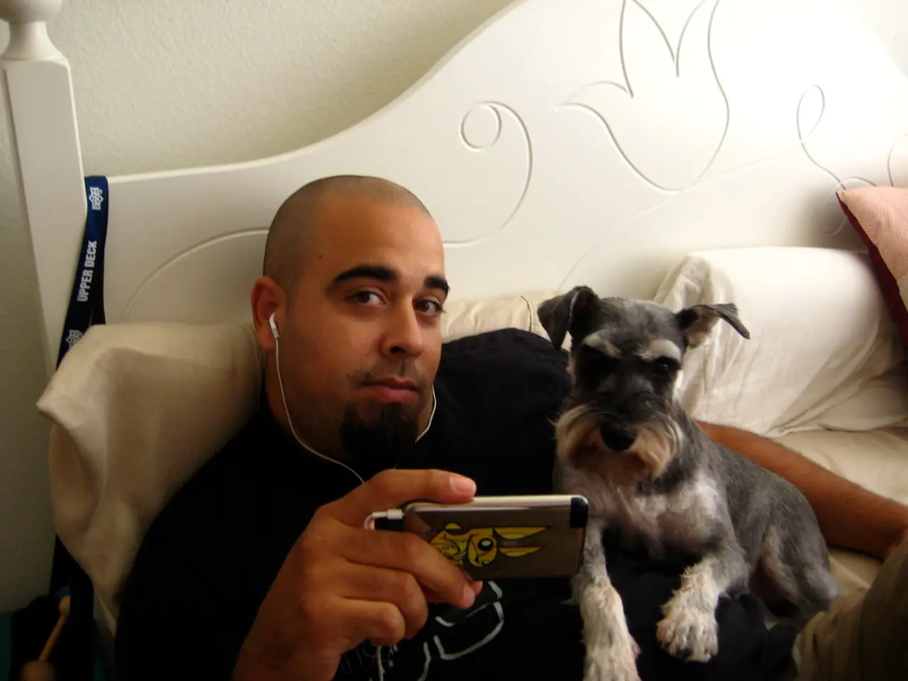 The picture is taken inside a room. One person is lying on the bed. He is holding one phone and a...