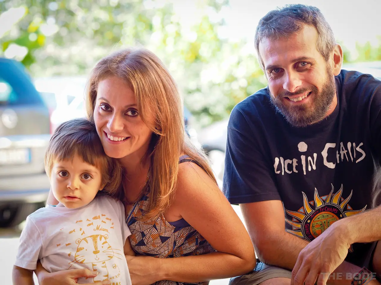 In this image in the front there are persons smiling. In the background there are cars and trees.