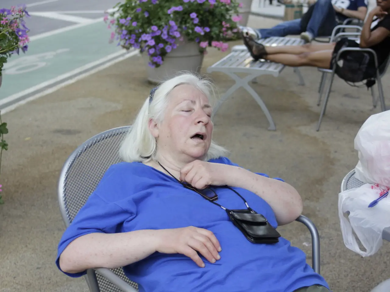 A woman is sitting on a chair and sleeping. There are other people at the back. There are flowers.