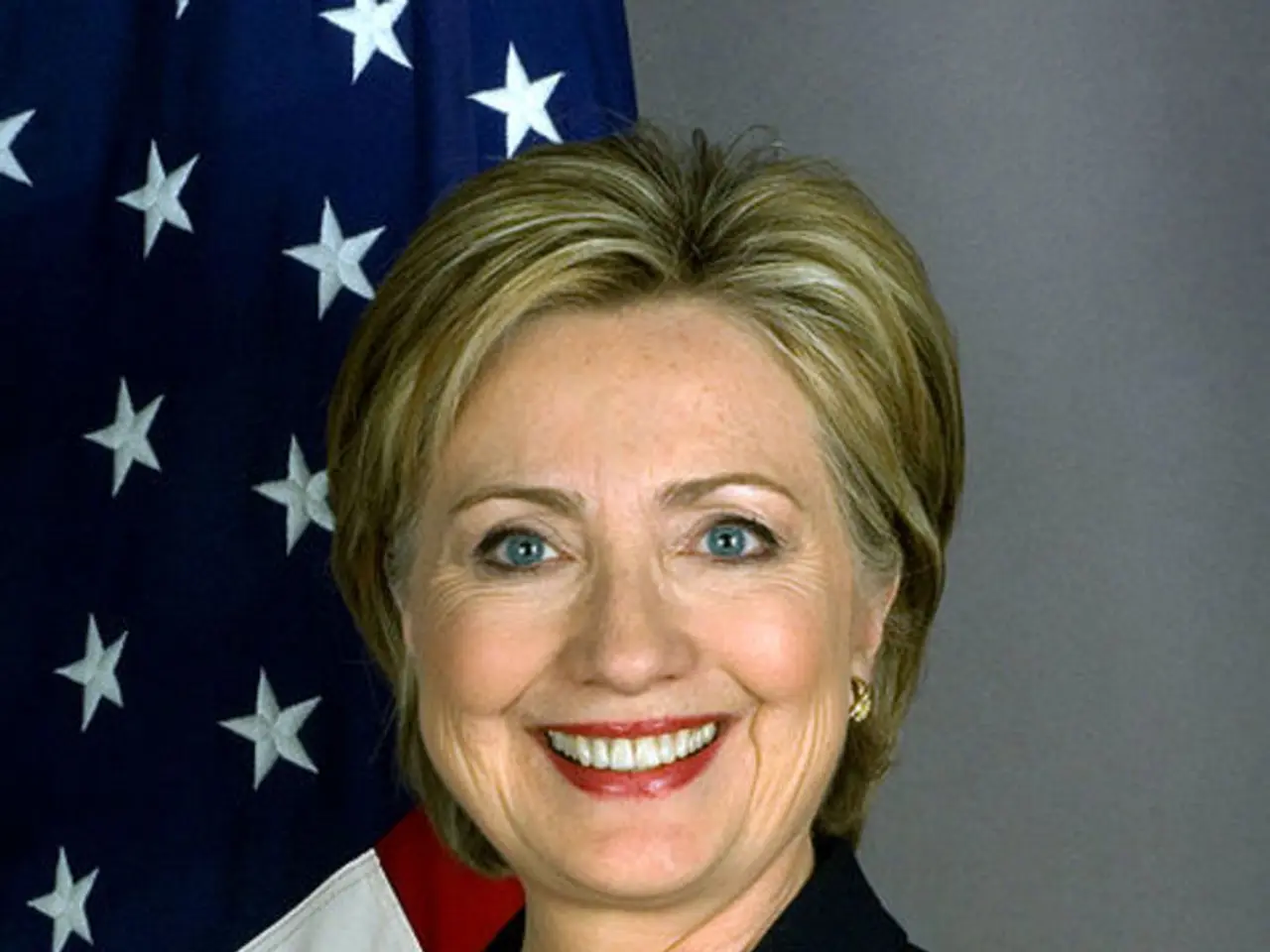 In this image we can see a woman smiling. In the background we can see the flag and also the wall.