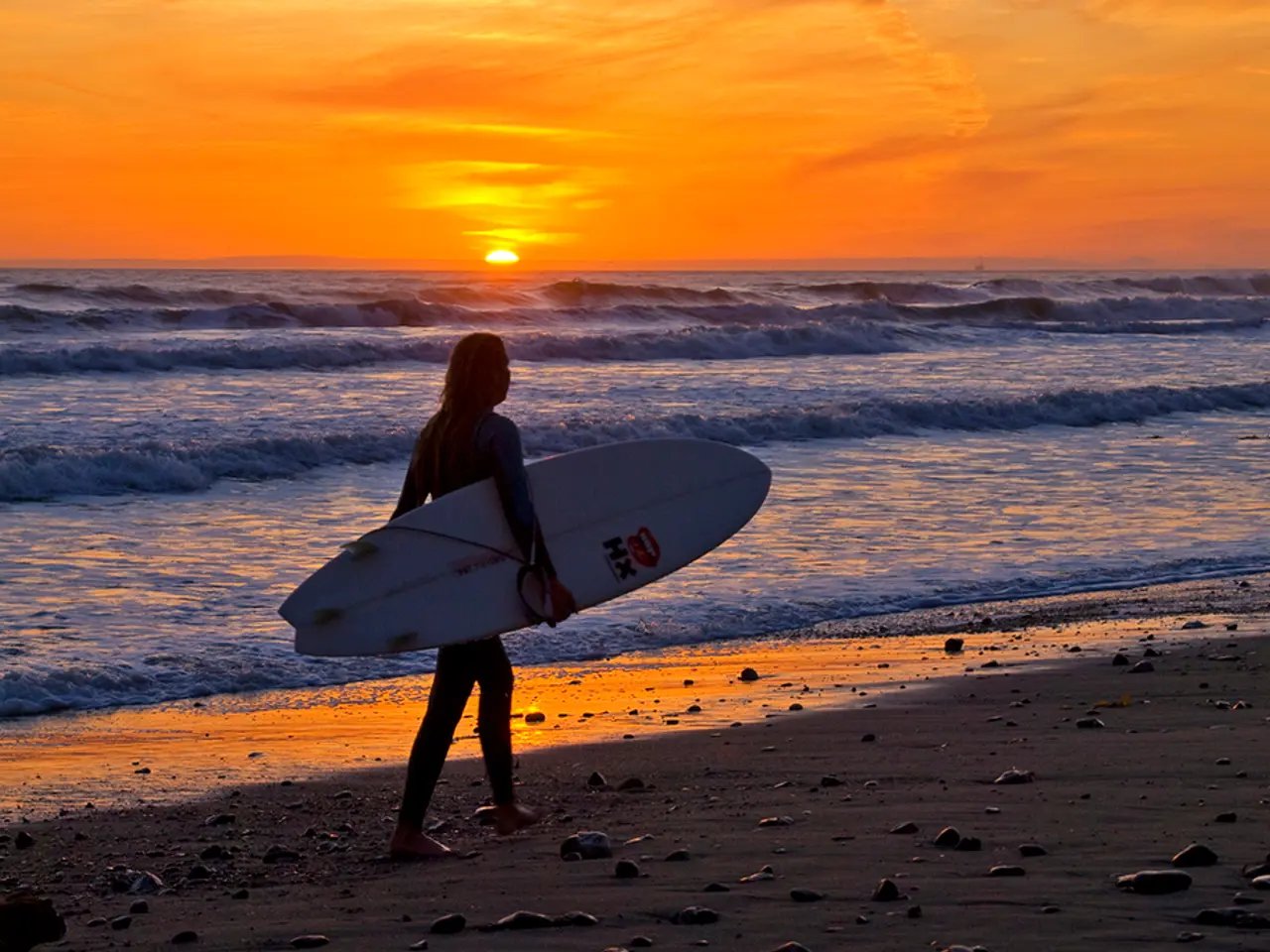 This picture is clicked at the beach during sunset. The woman in the image walking and holding a...