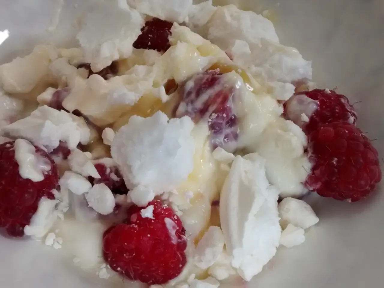 In this image there is a food item in the bowl. There are a few raspberries and there is cream.