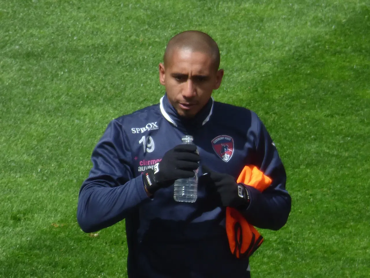The image shows a man standing on top of a lush green field, holding a bottle in one hand and an...