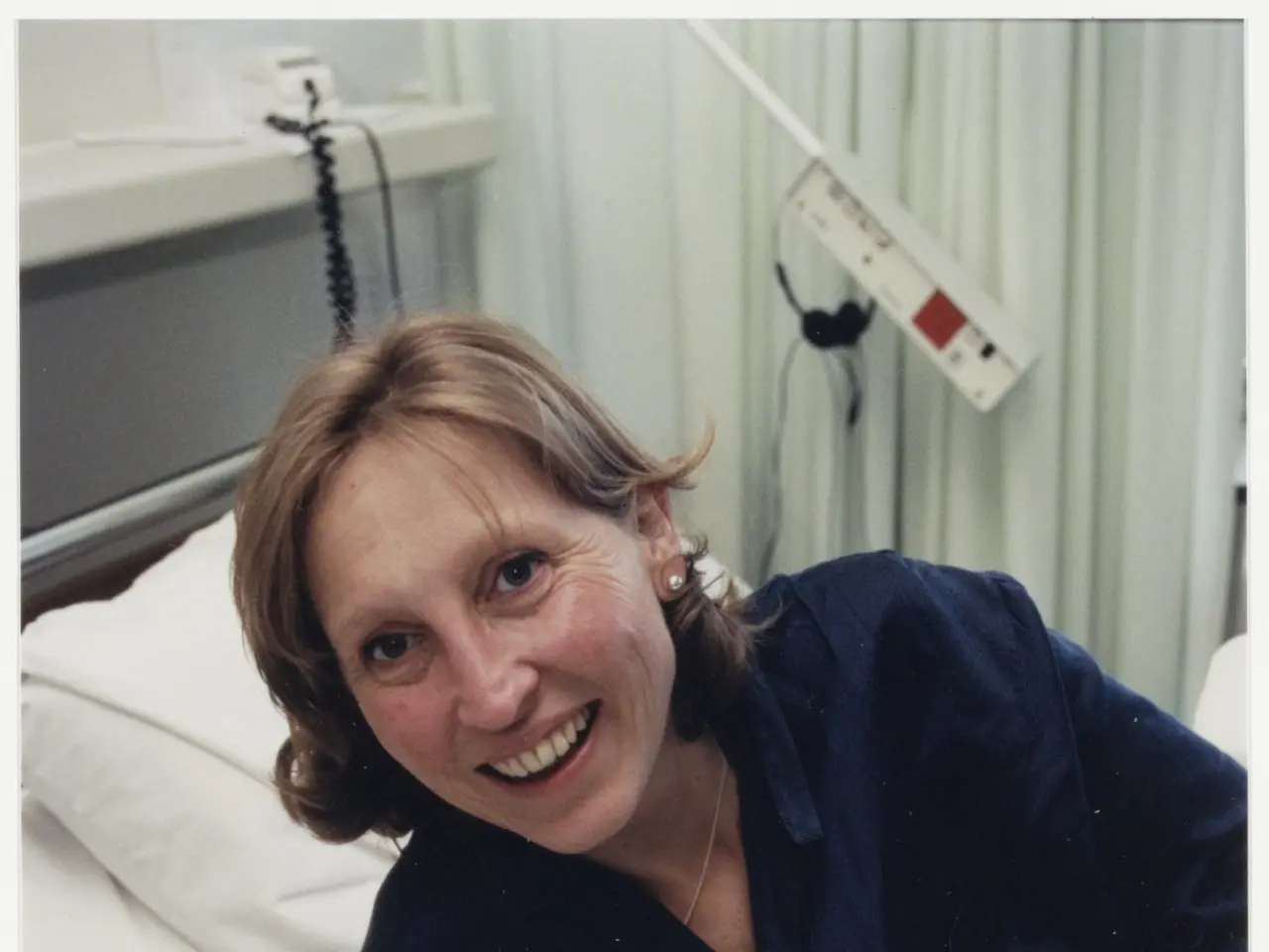 The image shows a woman in a hospital bed, cradling a baby in her arms. She is wearing a blue dress...