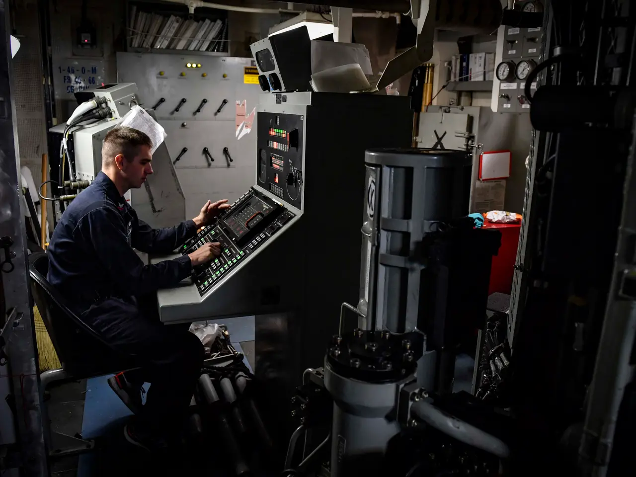 The image shows a man sitting at a desk in front of a computer, surrounded by various machines and...