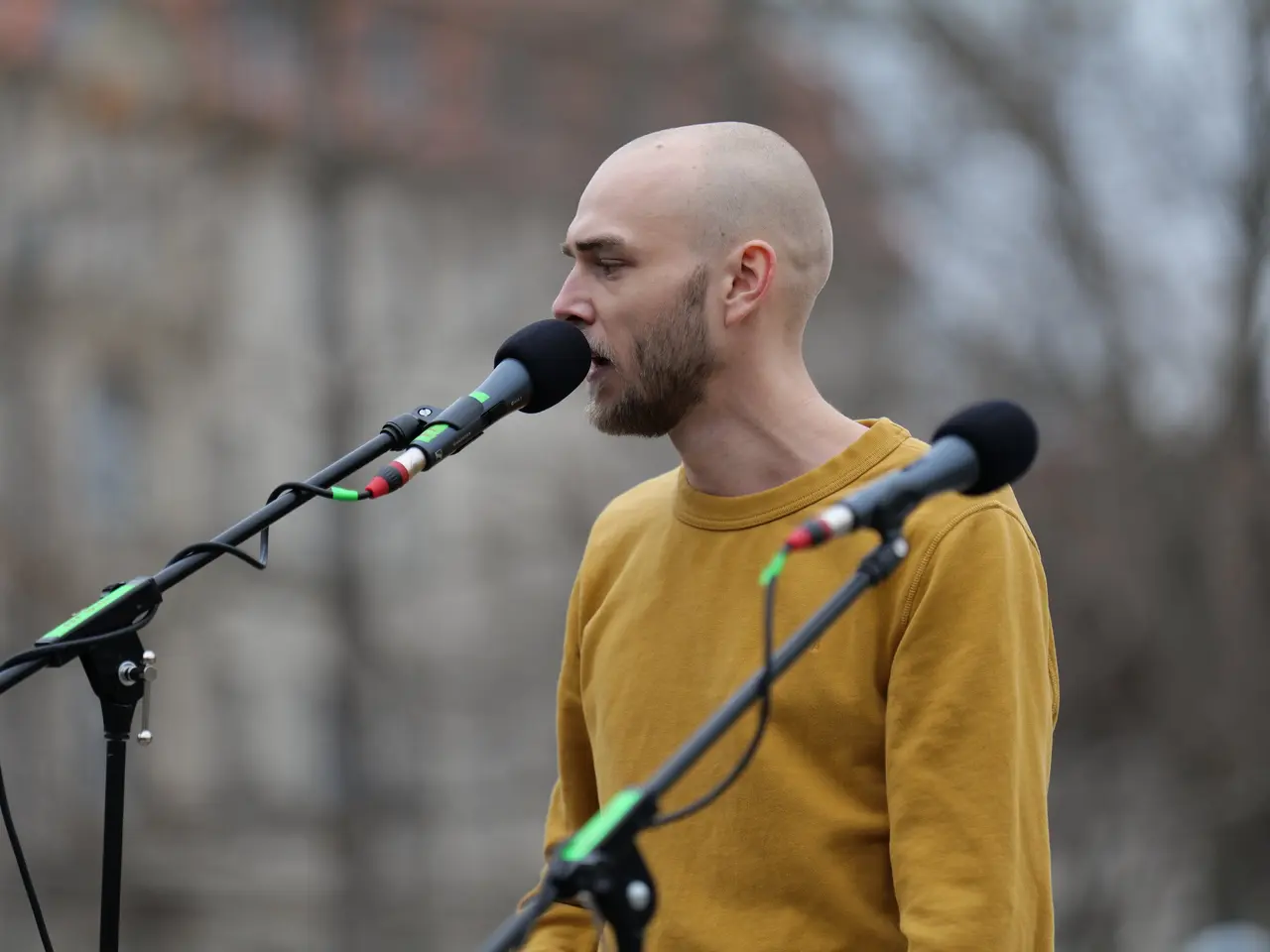 The image shows a bald man wearing a yellow t-shirt singing into a microphone in front of a blurred...