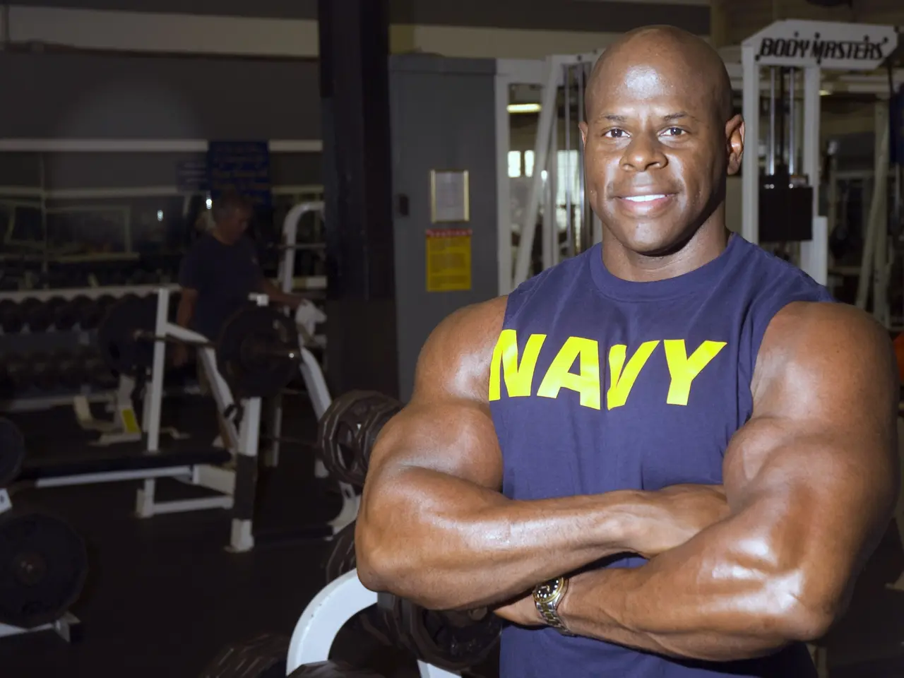 The image shows a bodybuilder standing in a gym with his arms crossed, wearing a blue t-shirt and...