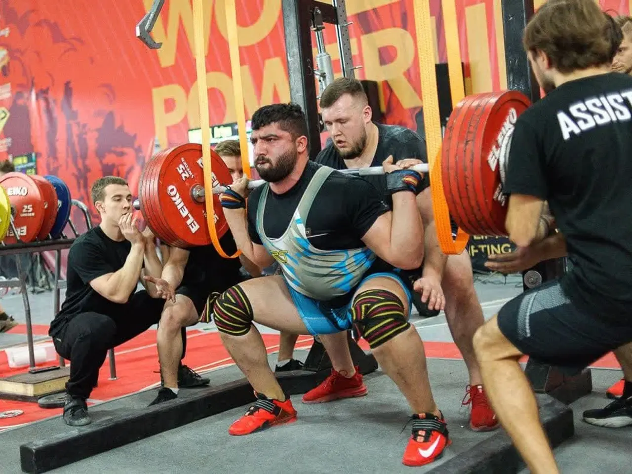 The image shows a group of men in a gym doing squats with barbells, surrounded by gym equipment and...