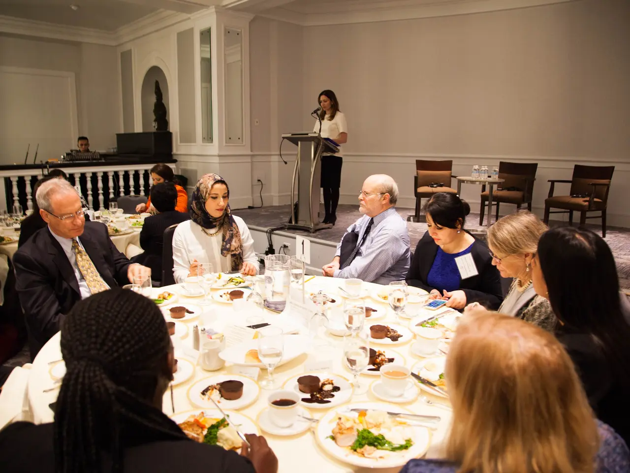 The image shows a group of people sitting around a table with plates of food, glasses, cups,...