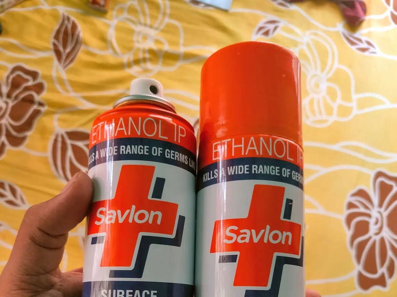The image shows a person holding two cans of Savlon surface disinfectant spray in their hand, with...