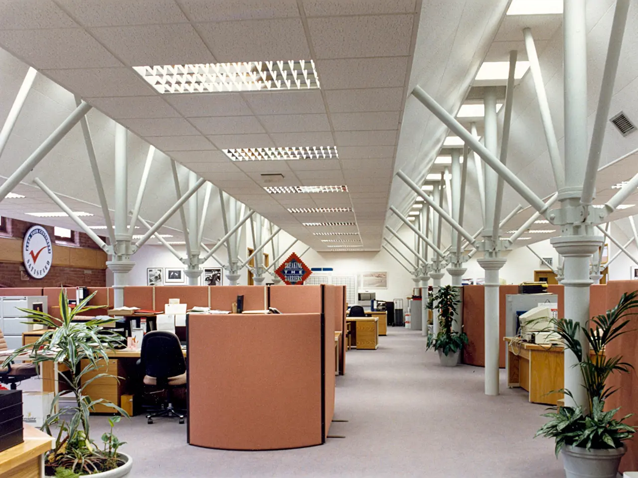 The image shows an empty office with lots of desks and chairs, houseplants, pillars, a clock on the...