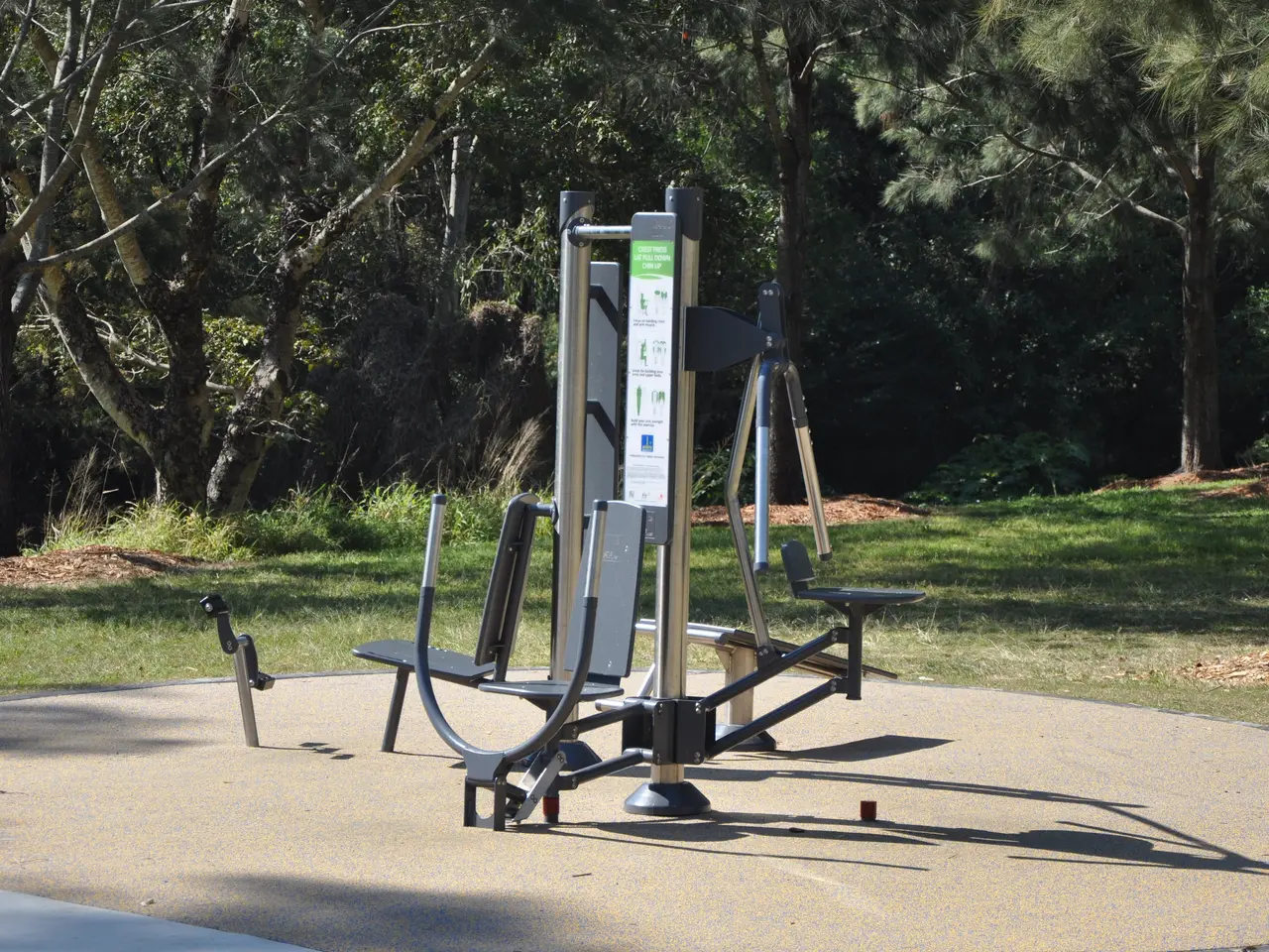 The image shows a park with a variety of exercise equipment in the middle of it, surrounded by lush...