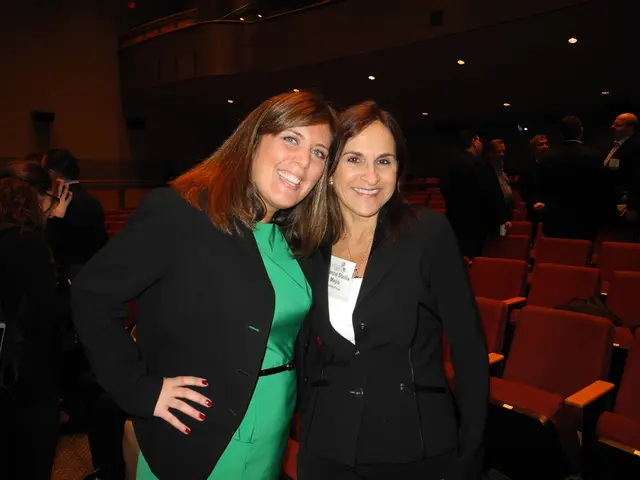 In this image there are two women wearing black coat standing in the front, smiling and giving and...