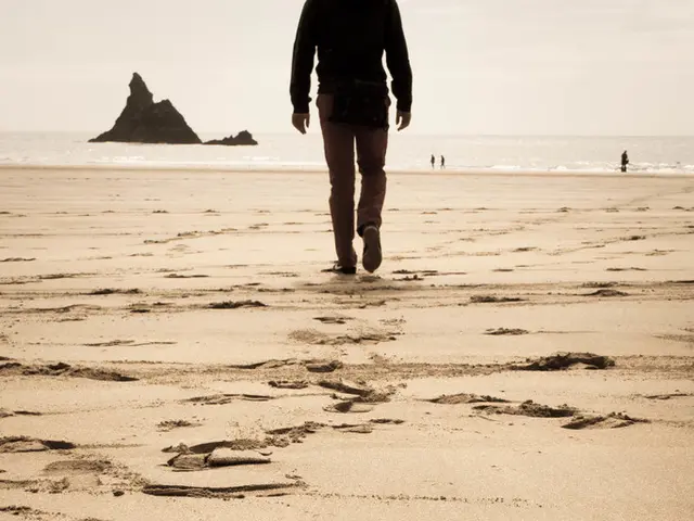 In the image there is a person walking on beach, in the front there is a small hill in the ocean...