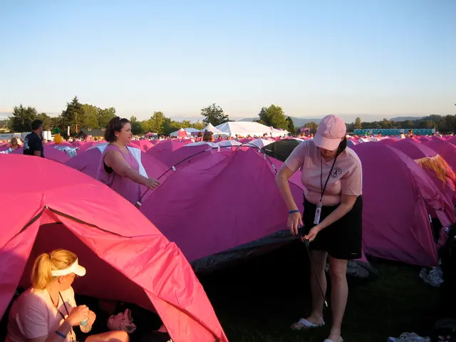 Here we can see tents and people. Far there is a house and trees. This woman is holding a bottle.