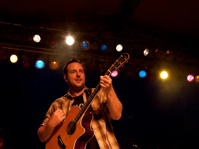 In the middle there is a man he wear check shirt , he is playing guitar, he is staring at...