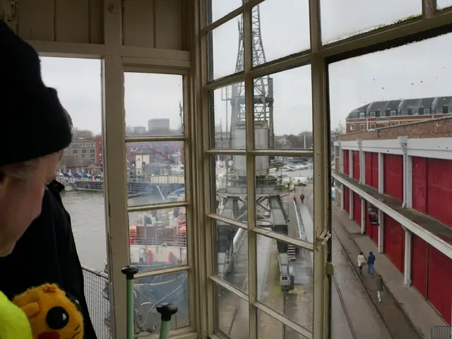 In this image there is a person operating a tower crane, in front of the crane there are buildings...