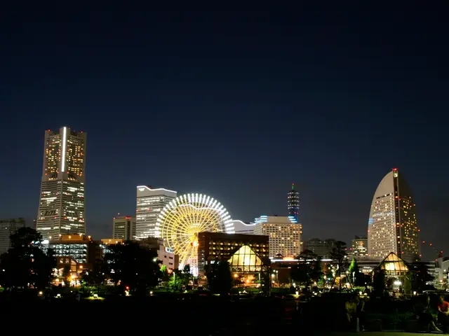 In this picture I can see few buildings with lighting and i can see few people seated and few trees...
