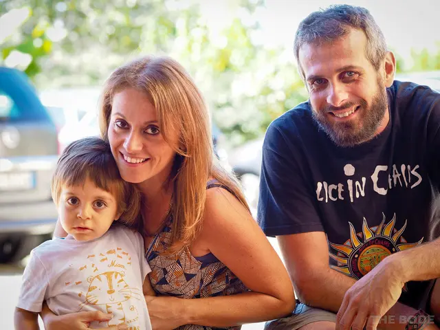 In this image in the front there are persons smiling. In the background there are cars and trees.