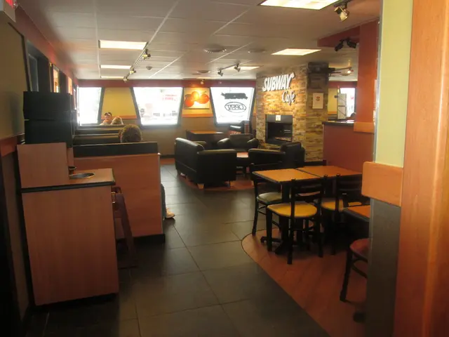 The picture is taken in a subway cafe where we can see number of tables,chairs and sofas. There are...