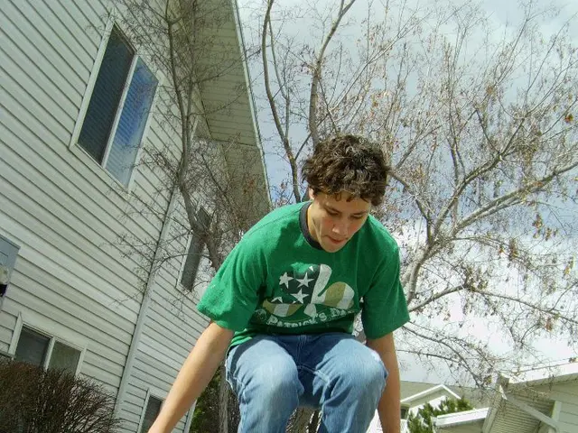 In this image we can see one man in green T-shirt humping, some houses, one small wall, some trees,...
