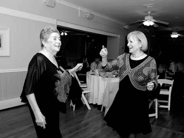 there are two women dancing and there are some people sitting on a chair with table in front of...