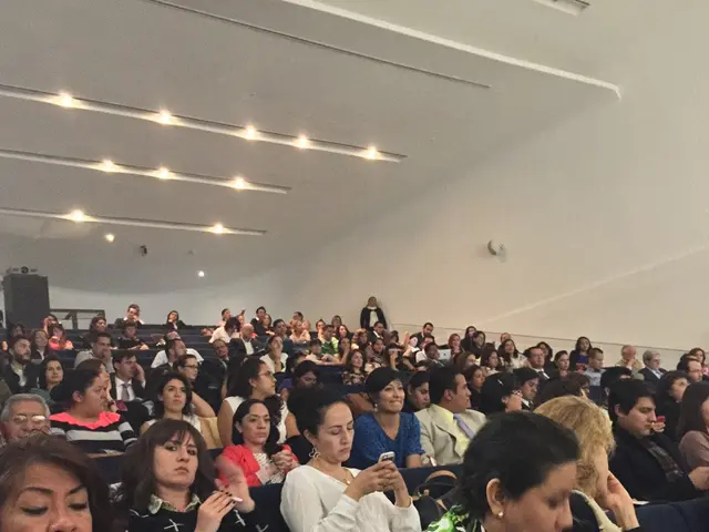 There are many people sitting in the room. In the ceiling there are lights. In the bottom left a...