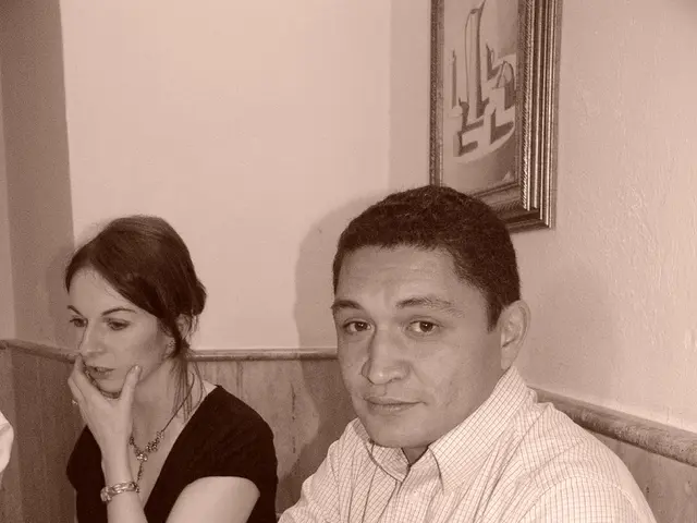 There is a man and woman sitting around the table. There is napkin and spoons and glass with water.