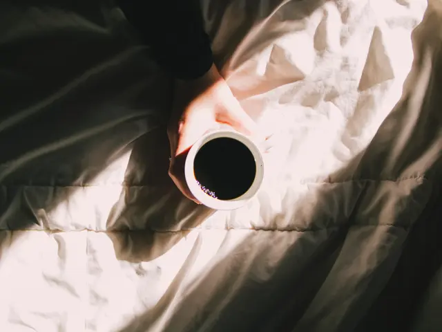 The image shows a person holding a cup of coffee in their hand, resting on a bed with a white cloth...