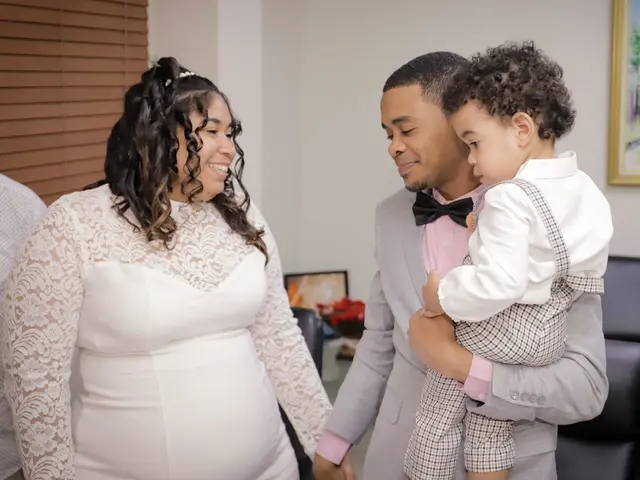 The image shows a pregnant woman in a white dress and a man in a suit holding a baby, standing in...