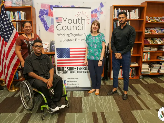 The image shows a group of people standing next to each other in front of bookshelves, with a man...
