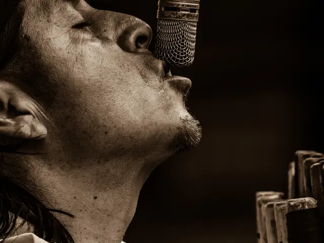 The image shows a man singing into a microphone in front of a dark background. He is wearing a...
