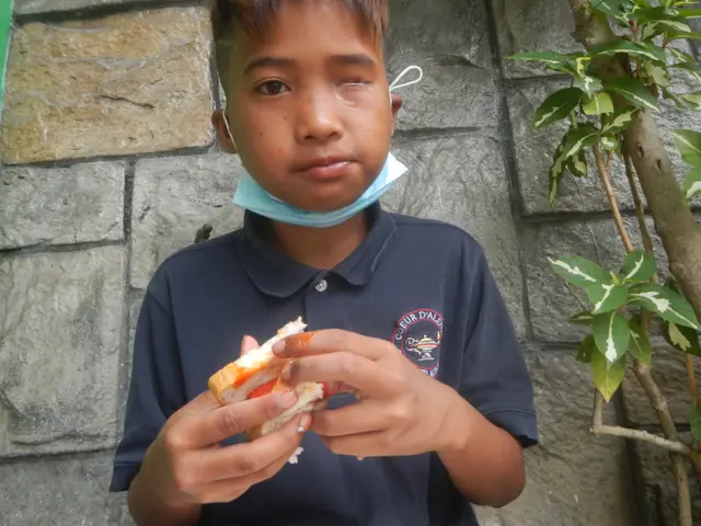 The image shows a young boy wearing a blue t-shirt and a face mask, holding a sandwich in his...