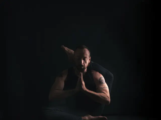 The image shows a man sitting in a yoga pose on a black mat, with a dark background. He is wearing...