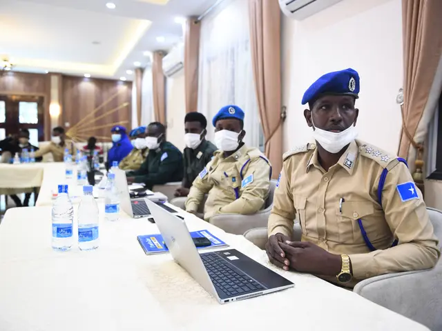The image shows a group of police officers sitting around a table with laptops, bottles, books, and...