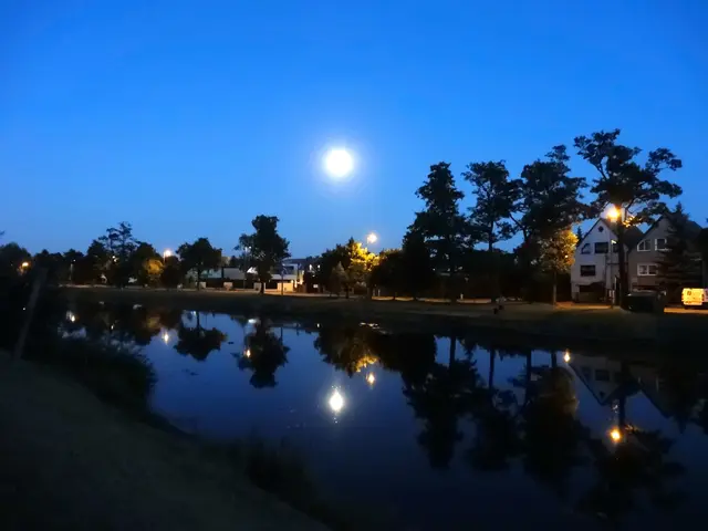 The image shows a full moon rising over a pond in the middle of a park at night. The pond is...
