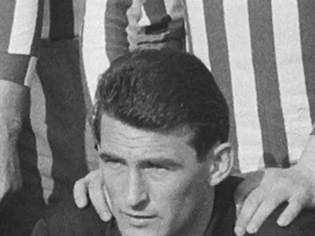 The image shows a black and white photo of a man sitting on top of a soccer field, wearing a...