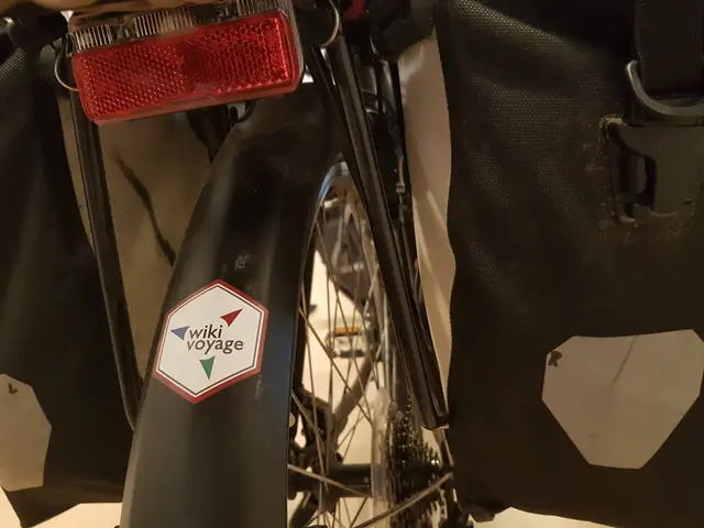 The image shows a close up of a bike with a bag on the back of it. The bike has a wheel and a...