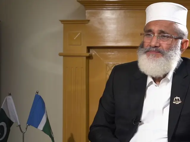 The image shows a man in a suit and hat sitting at a table with two flags in front of him. He is...