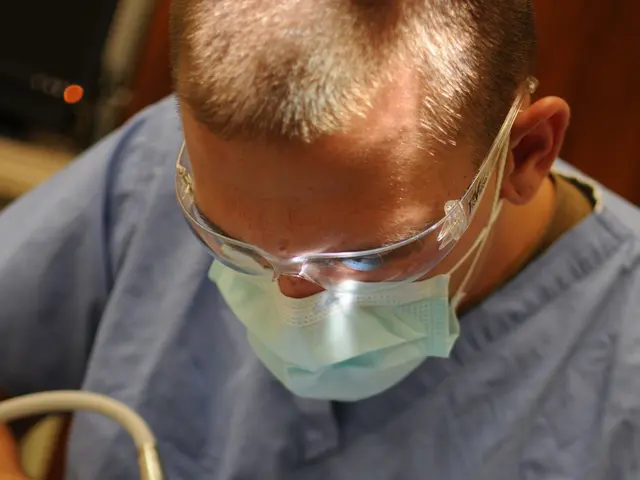 The image shows a man lying on a chair while a dentist wearing glasses, a mask, and gloves is...