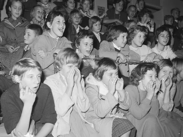 The image shows a black and white photo of a group of children sitting in front of a crowd, with a...