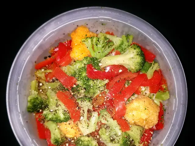 The image shows a plastic container filled with broccoli and red peppers on top of a table. The...