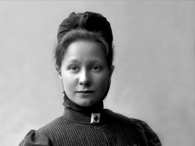 The image shows a black and white photo of a woman in a nurse's uniform, sitting on a chair. She...