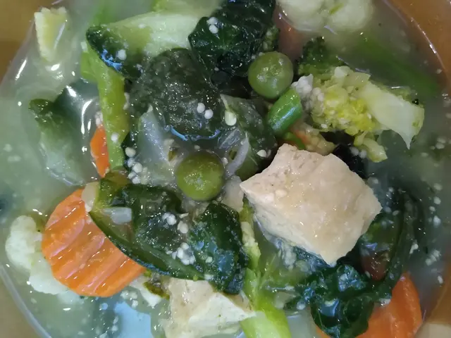 The image shows a bowl of soup with tofu and vegetables in it, sitting on a table. The soup is a...
