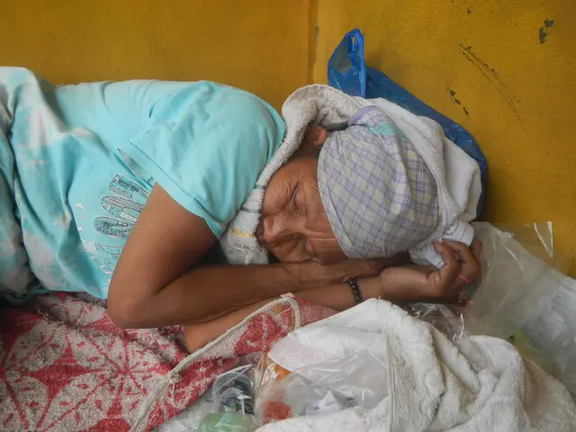 The image shows an elderly woman sleeping on a bed with a blanket draped over her head, surrounded...