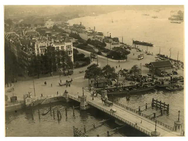 The image shows a black and white photo of a city with a bridge spanning a body of water, with a...