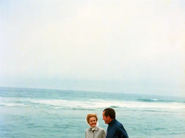 The image shows a man and woman walking on the beach, with the sea and sky in the background. They...