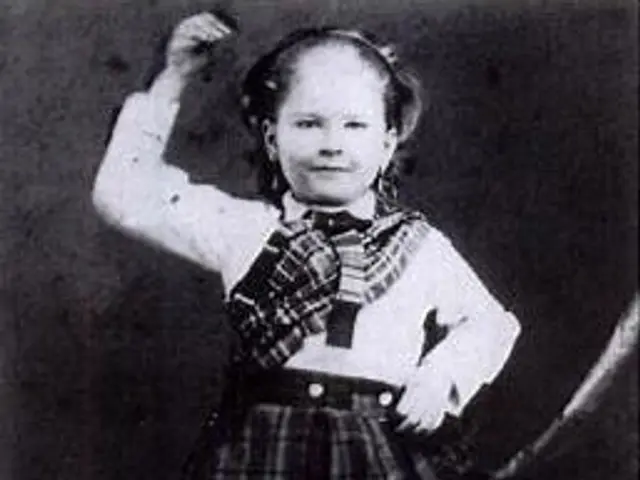 The image shows an old black and white photo of a young girl in a school uniform, standing on the...